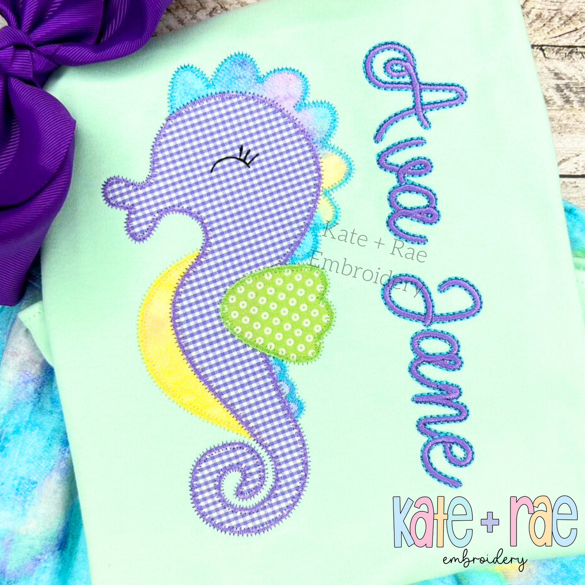 Delilah Outline Satin Stitch Embroidery Font: The name Ava Jane in purple outline script beside a gingham seahorse applique on a mint shirt
