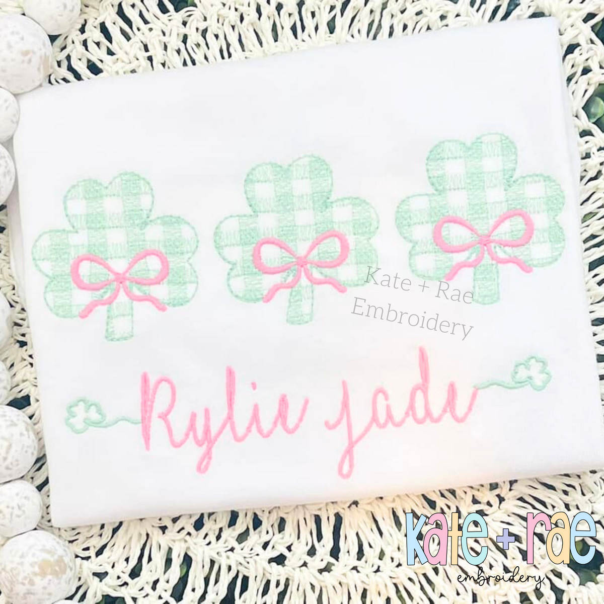 Plaid Shamrock Trio with Bows Sketch Stitch Embroidery Design: Three green gingham shamrocks with pink bows above the name Rylie Jade in pink script on a white shirt