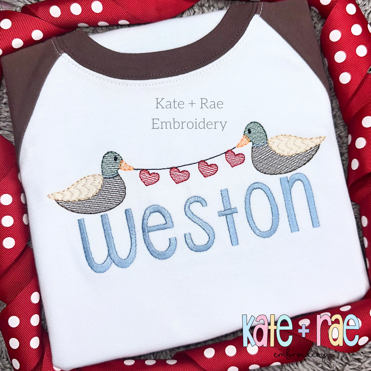 Mallard Duck with Heart String Sketch Stitch Embroidery Design: Two mallard ducks holding a string of pink hearts above the name weston in blue on a white shirt