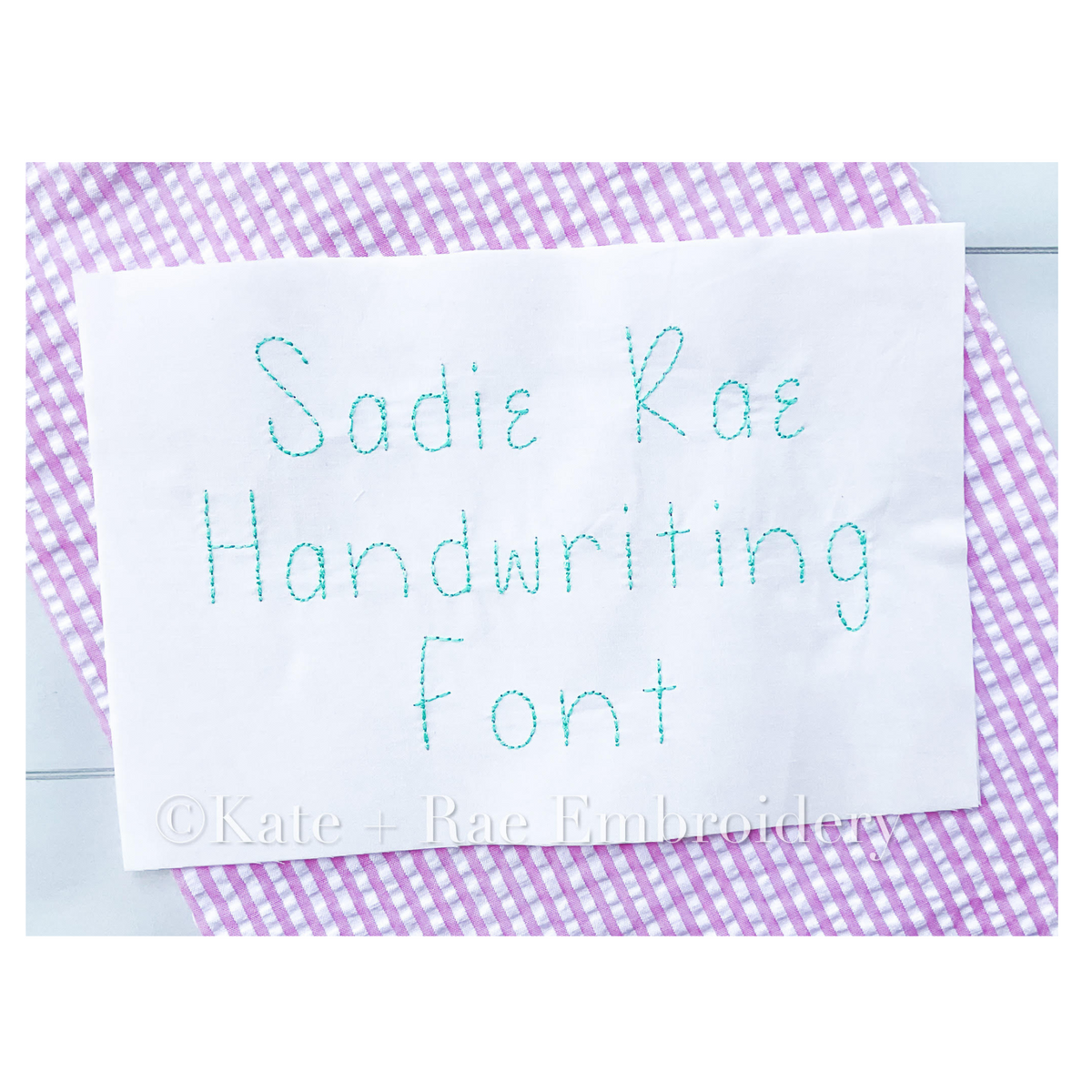 Sadie Rae Handwriting Embroidery Font: The words Sadie Rae Handwriting Font stitched in mint thread on white fabric over purple gingham