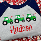 Heart Tractor Trio Fill Stitch Embroidery Design: Three green tractors with hearts above the name Hudson in red on a gray and navy raglan shirt