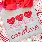 Heart Sucker Trio with Bows Satin Stitch Applique Embroidery Design: Three red polka dot heart sucker appliques with pink bows above the name caroline in red script on a gray sweatshirt