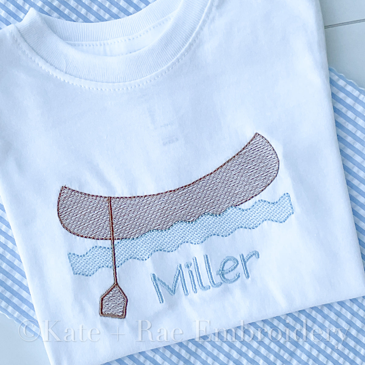 Canoe Sketch Stitch Embroidery Design: Brown sketch stitch canoe with a paddle over blue waves above the name Miller stitched on a white shirt