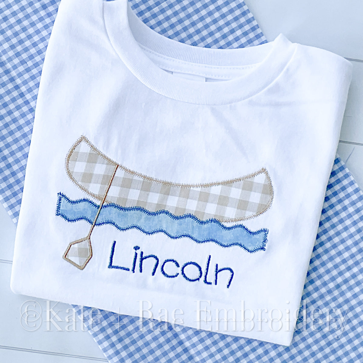 Canoe Zigzag Applique Embroidery Design: Tan gingham canoe with a paddle over blue waves above the name Lincoln stitched on a white shirt