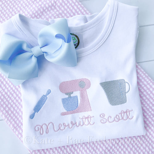 Baking Mixer Trio Fill Stitch Embroidery Design: Rolling pin, pink stand mixer, and measuring cup stitched above the name Merritt Scott on a white shirt with a blue bow