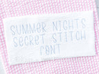 Summer Nights Secret Stitch Embroidery Font: The words Summer Nights Secret Stitch Font stitched in light gray on white fabric over pink gingham