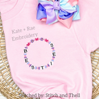 Friendship Embroidery Font: Friendship bracelet circle of beaded letters spelling besties on a pink shirt with a purple bow