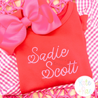 Valentine Heart Satin Stitch Embroidery Font: The name Sadie Scott in pink heart satin script on a red shirt