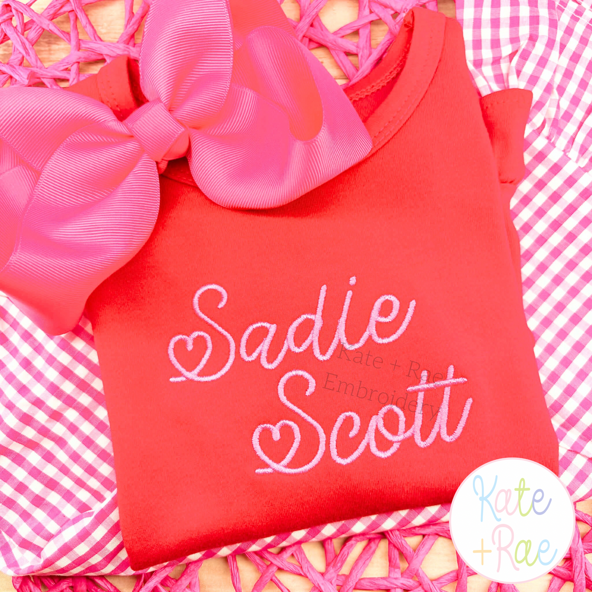 Valentine Heart Satin Stitch Embroidery Font: The name Sadie Scott in pink heart satin script on a red shirt