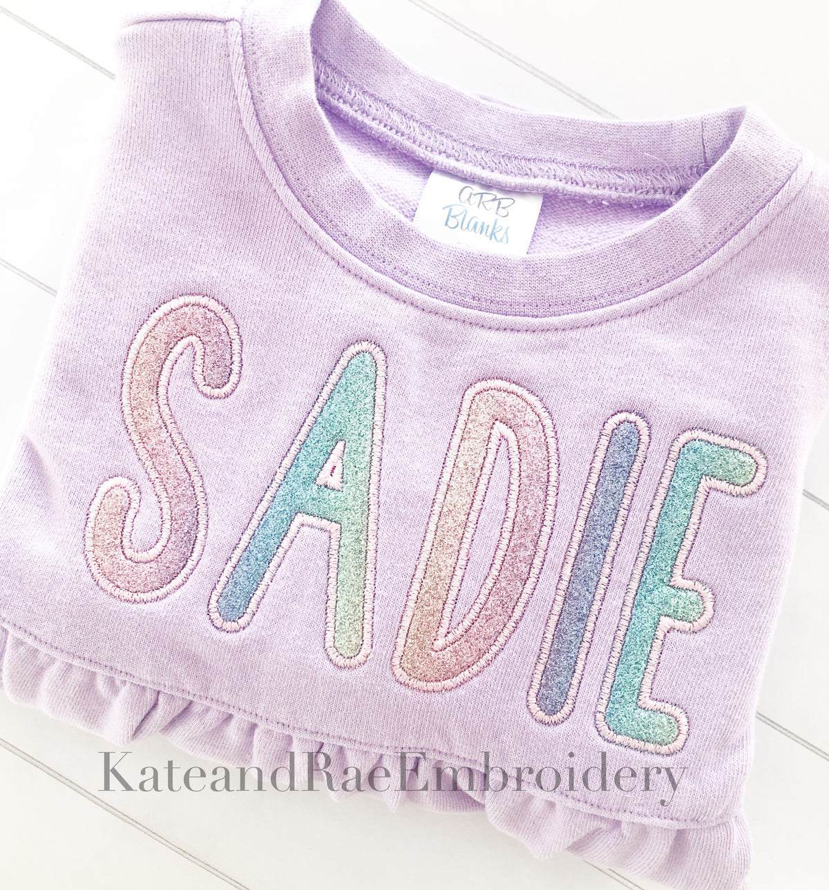 Simple Life Satin Stitch Applique Embroidery Font: The name Sadie in pastel glitter satin applique letters on a lavender sweatshirt