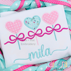 Heart Sucker Trio with Bows Satin Stitch Applique Embroidery Design: Three patterned heart sucker appliques with pink bows above the name mila in aqua script on a white shirt