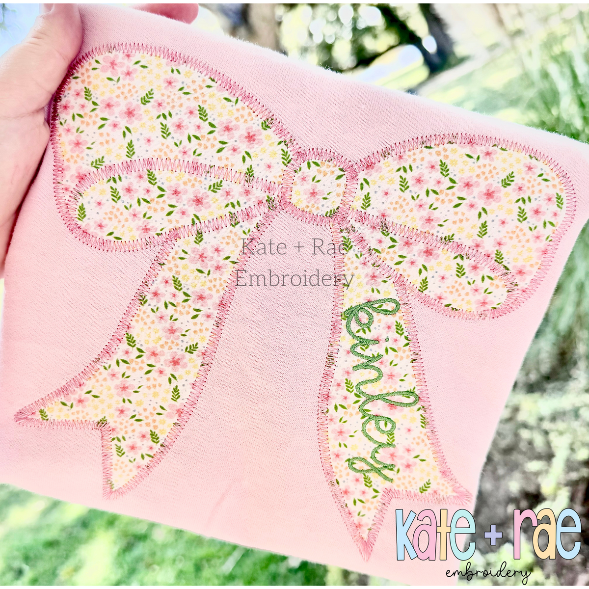 Kinley Bow Zigzag Stitch Applique Embroidery Design: Large pink floral bow applique with the name kinley on the tail on a pink shirt