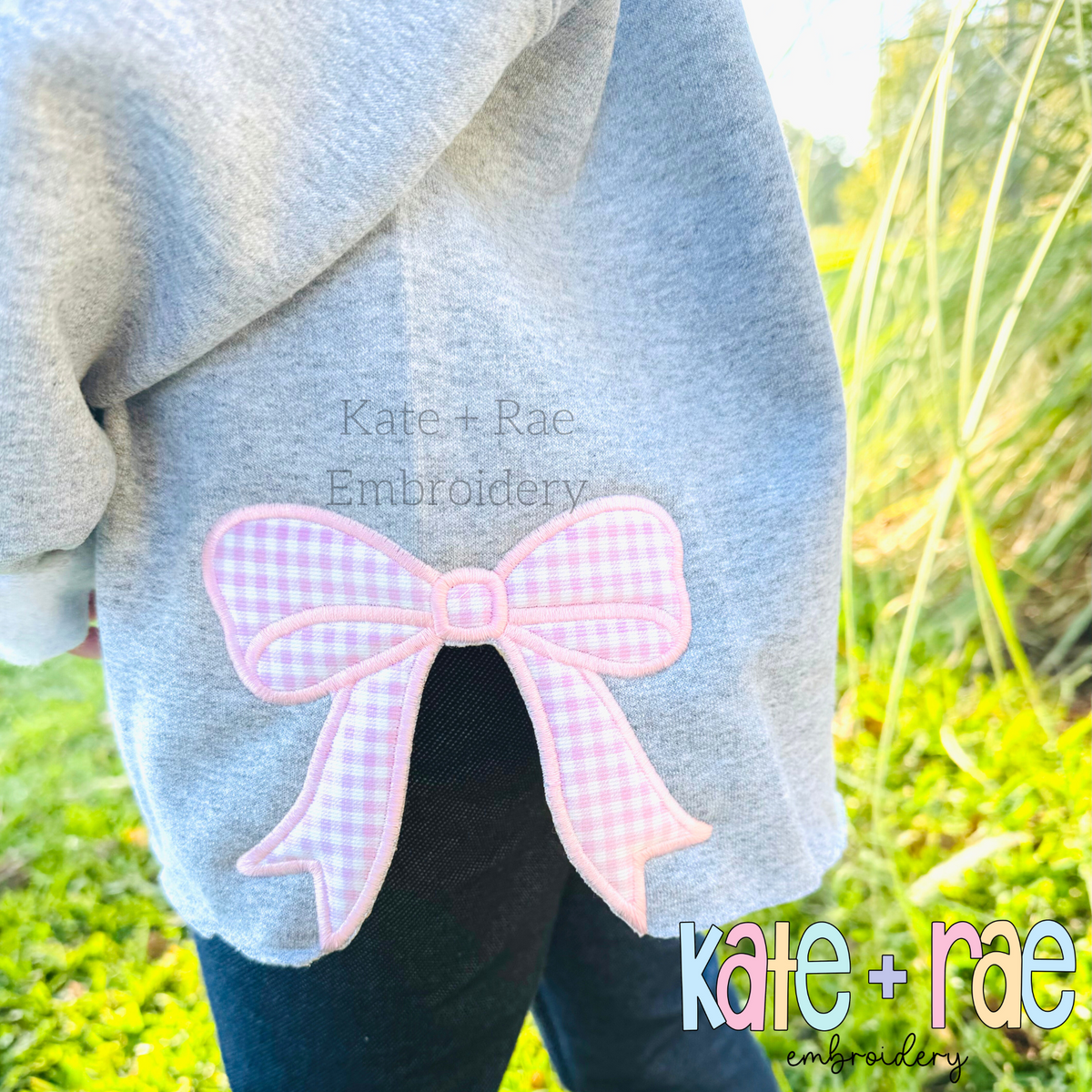 Kinley Bow Satin Stitch Applique Embroidery Design: Pink gingham bow applique stitched over the side slit of a gray sweatshirt