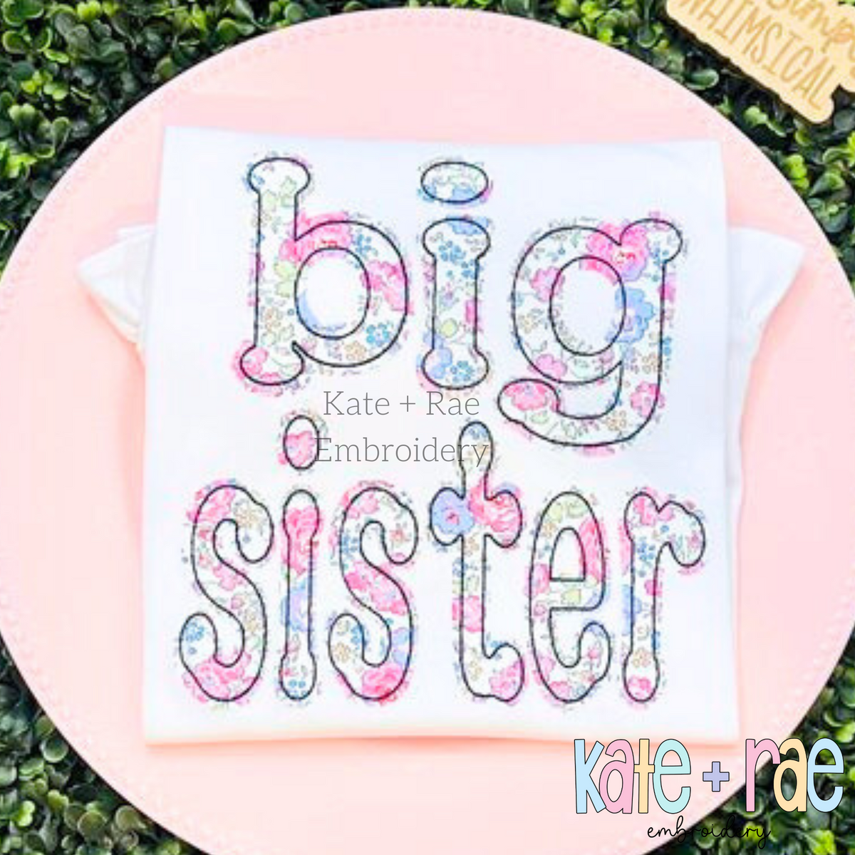 Dottie Bean Stitch Applique Embroidery Font: The words big sister in pastel floral applique letters on a white shirt