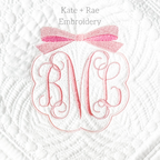 Scalloped Monogram Frame with Bow Embroidery Design: Pink scalloped frame with a pink bow around a script monogram on a white quilted blanket