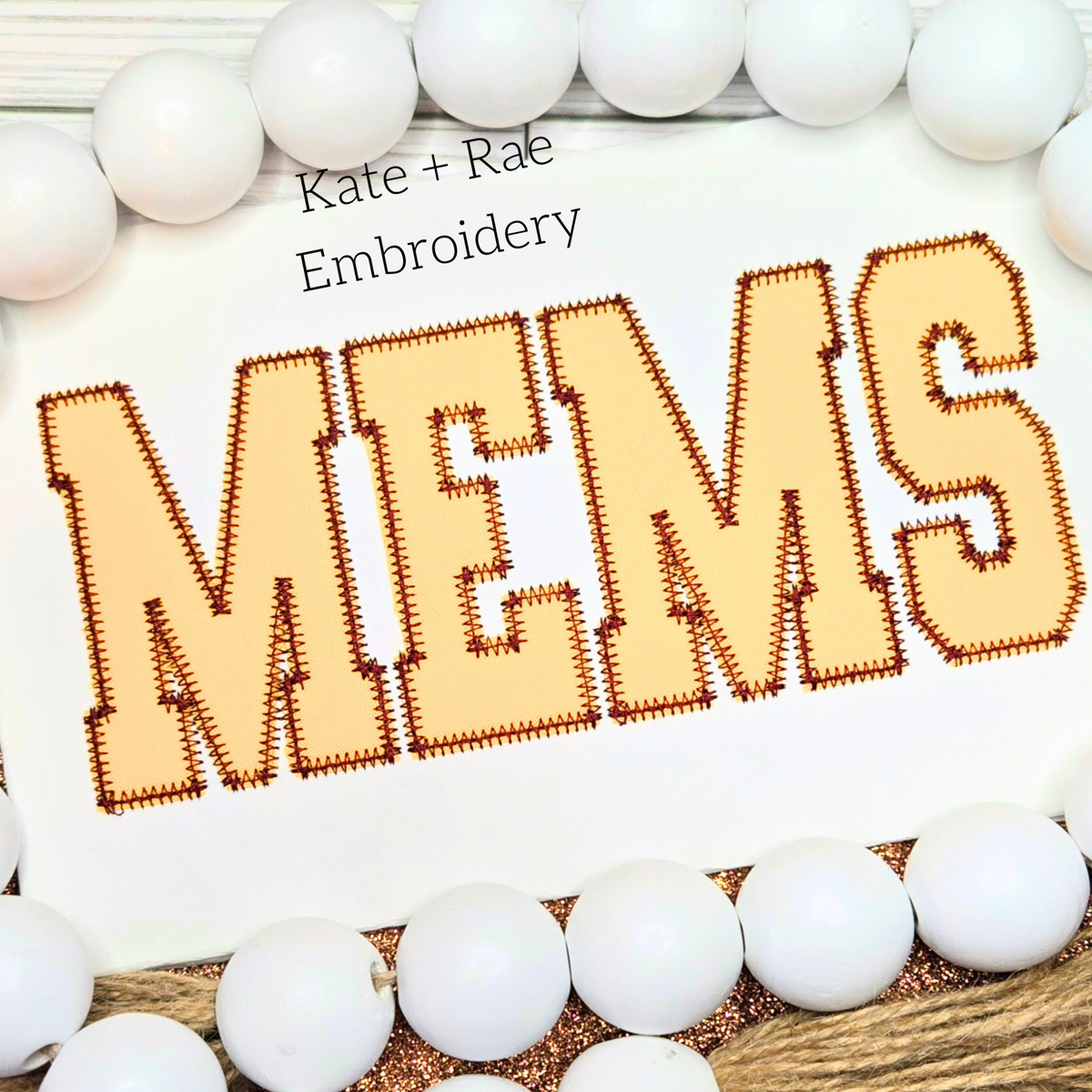 Varsity Zigzag Stitch Applique Embroidery Font: The word MEMS in tan collegiate zigzag applique letters on white fabric