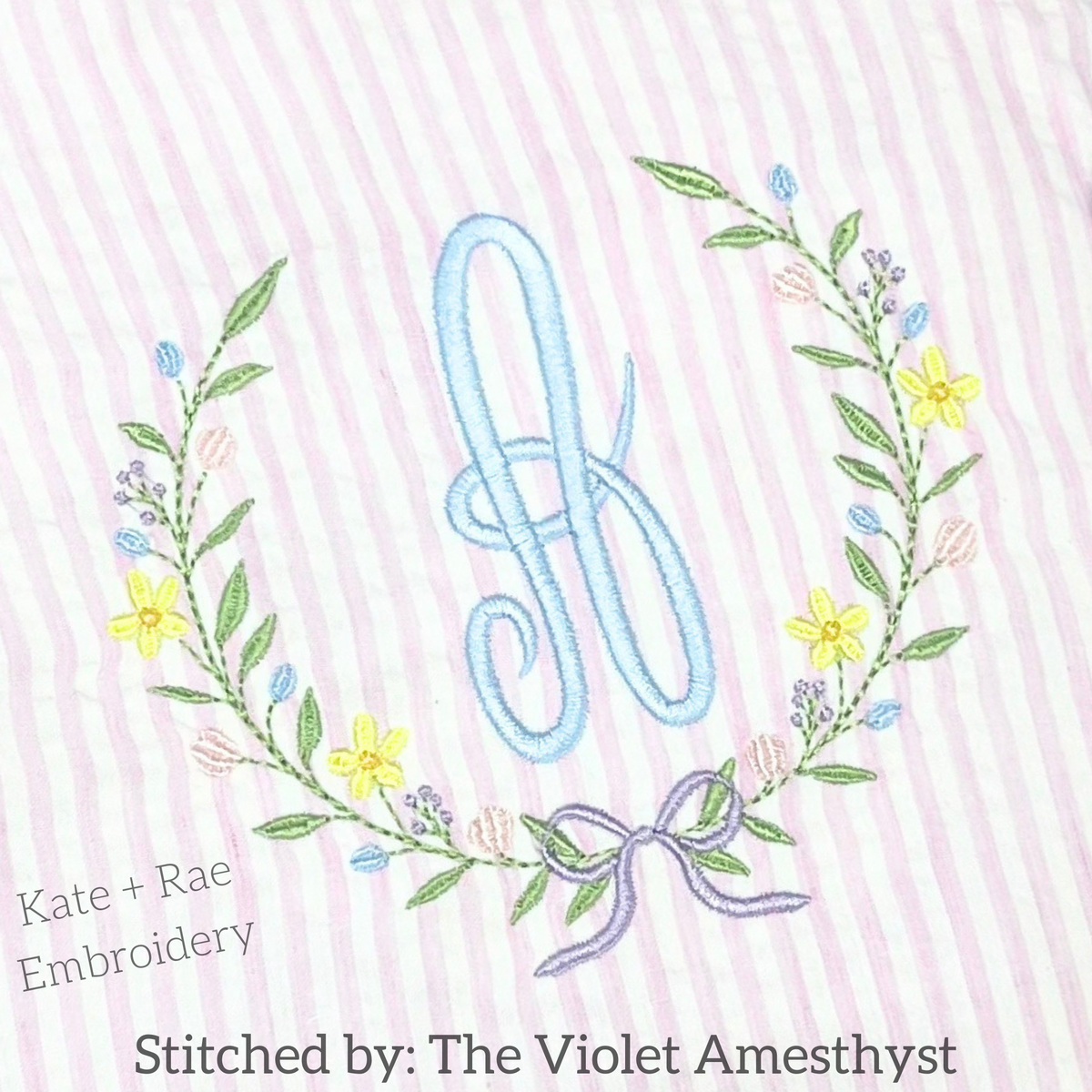 Wildflower Frame Fill Stitch Embroidery Design: Wildflower wreath with a purple bow around a blue script monogram on pink seersucker