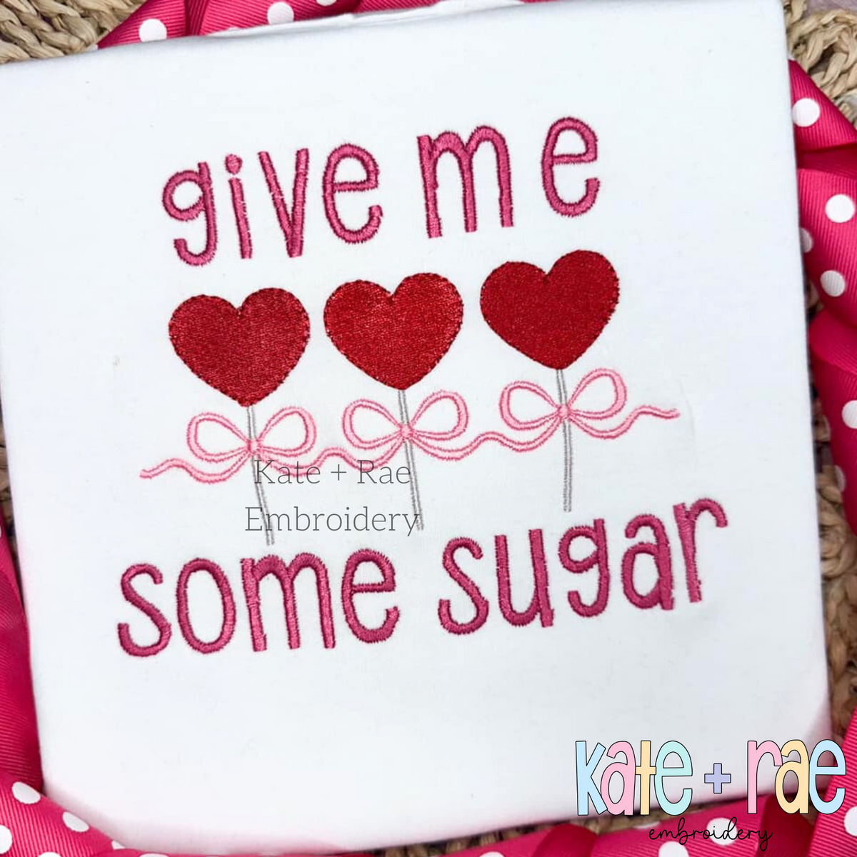 Heart Sucker Trio with Bows Fill Stitch Embroidery Design: The words give me some sugar in pink around three red heart suckers with a pink bow on a white shirt