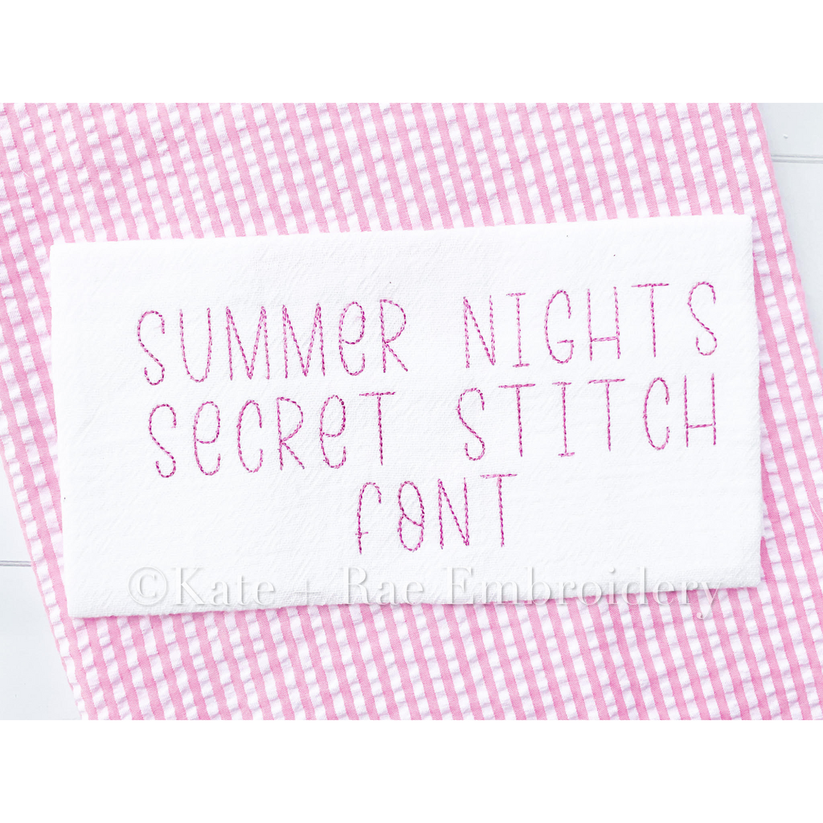 Summer Nights Secret Stitch Embroidery Font: The words Summer Nights Secret Stitch Font stitched in pink on white fabric over pink gingham