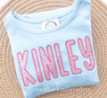 Simple Life Zigzag Stitch Applique Embroidery Font: The name Kinley in pink glitter applique letters on a light blue shirt
