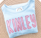 Simple Life Zigzag Stitch Applique Embroidery Font: The name Kinley in pink glitter applique letters on a light blue shirt