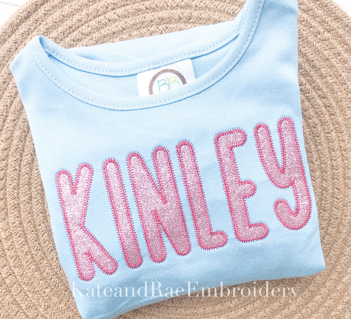 Simple Life Zigzag Stitch Applique Embroidery Font: The name Kinley in pink glitter applique letters on a light blue shirt