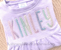 Simple Life Satin Stitch Applique Embroidery Font: The name Kinley in pastel glitter applique letters on a lavender ruffled sweatshirt